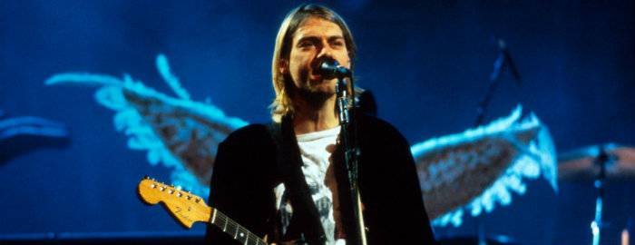 Kurt Cobain toca guitarra. Ao fundo a arte do disco In utero