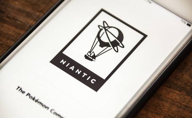 Fotografia de um celular com o logo da Niantic, desenvolvedora de Pokémon GO. O logo da empresa está na cor preta e possui um balão, o fundo da tela é branca. 